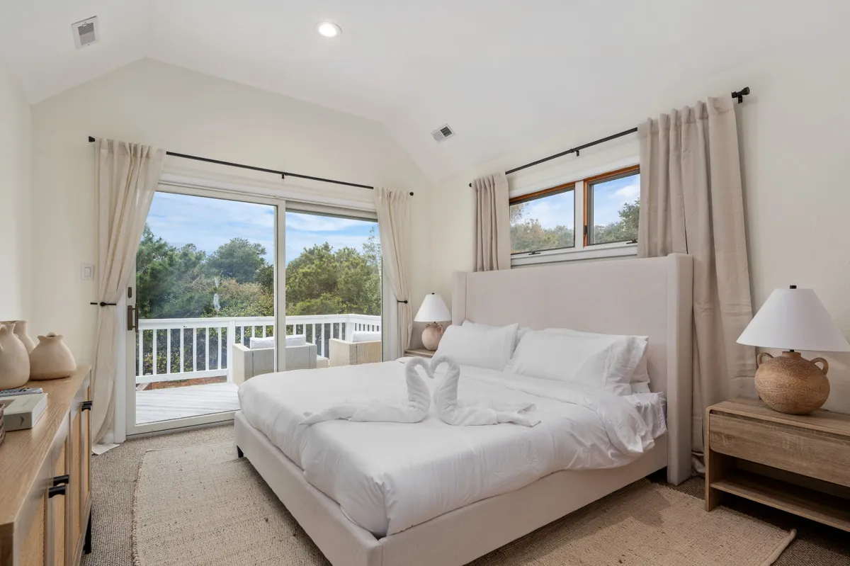 A serene bedroom featuring a comfortable bed positioned near a sliding glass door and large window, inviting in natural light. A stylish nightstand with a lamp adds to the tranquil atmosphere, while the room’s decor emphasizes relaxation and simplicity.
