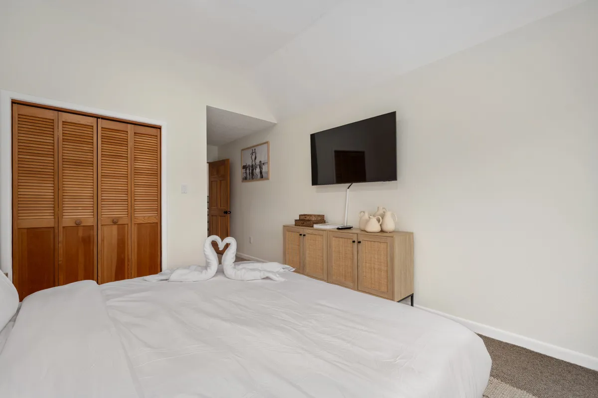 A tranquil bedroom with a neatly made bed adorned with elegantly arranged towels. The warm wooden storage unit and flat-screen TV suggest modern comforts, while a print on the wall adds subtle decor. The overall feel is both relaxing and luxurious.