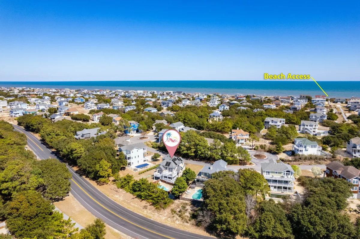 This image features a beach access sign in the foreground, with a prominent house to one side, lush greenery, and the ocean visible in the background. The scene provides a direct visual cue to the proximity and accessibility of Corolla’s beaches, reinforced by the inviting pathway leading to the shoreline.