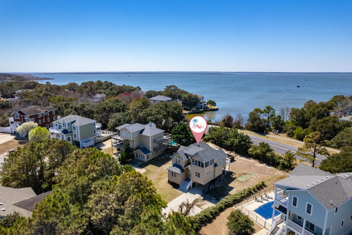 The scene features a tranquil waterfront with the house in view, nearby trees, and a glimpse of neighboring properties. The atmosphere is calm and inviting, suggesting opportunities for waterfront walks, relaxation, and proximity to local gathering spots.