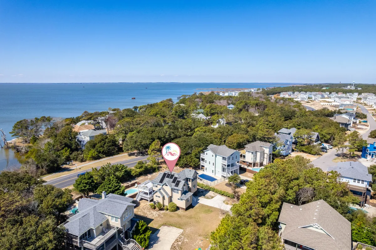 An elevated view looking out over an oceanfront neighborhood, with the ocean stretching into the horizon, tree-lined roads, and several houses visible. The scene captures a tranquil beach town atmosphere, illustrating both the community layout and the natural beauty of the coast.