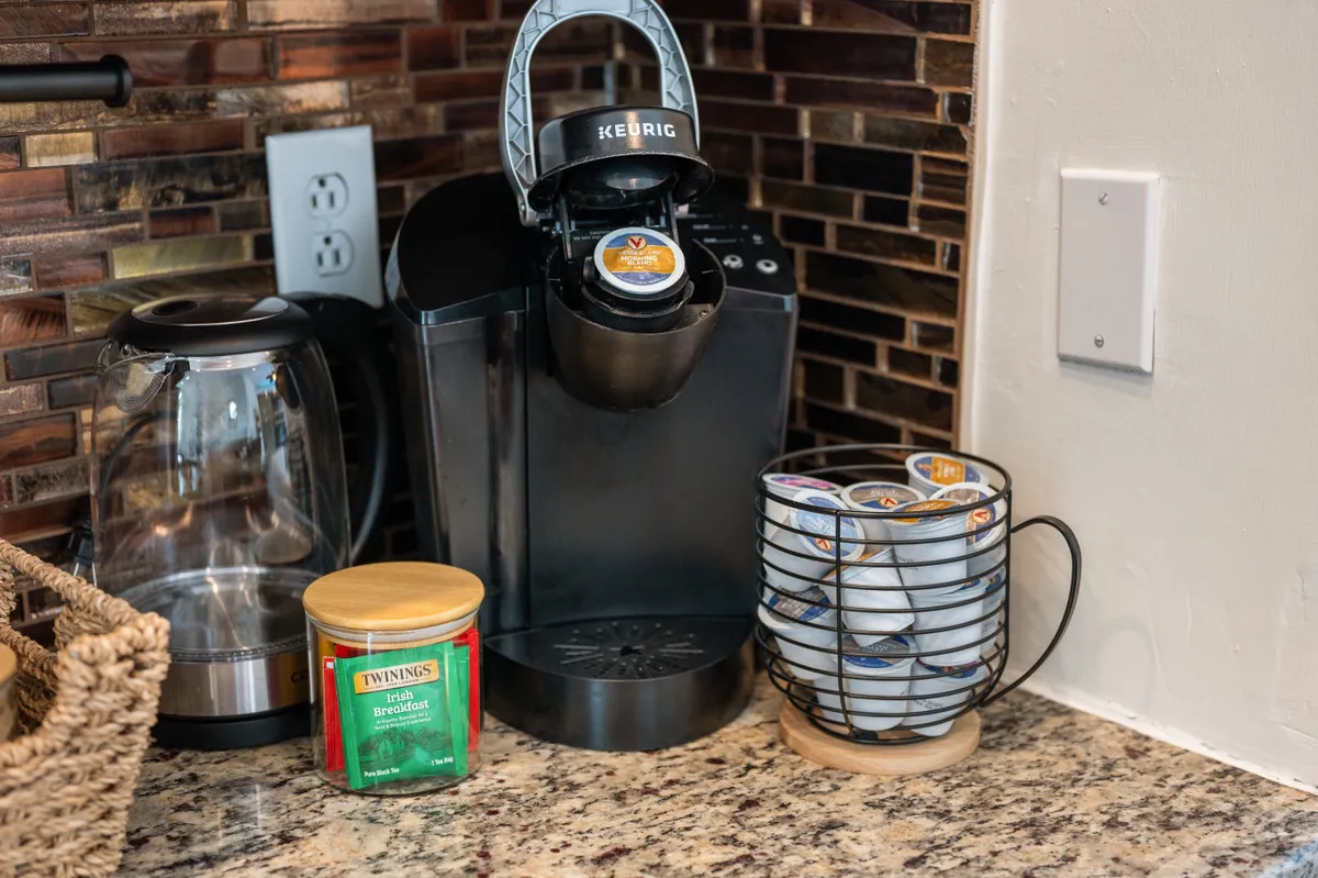 A close-up of the kitchen beverage station showcases a Keurig coffee maker, glass kettle, tea canister, and well-organized K-Cup storage. The setup is tidy and inviting, emphasizing both convenience and the small pleasures of a morning routine.