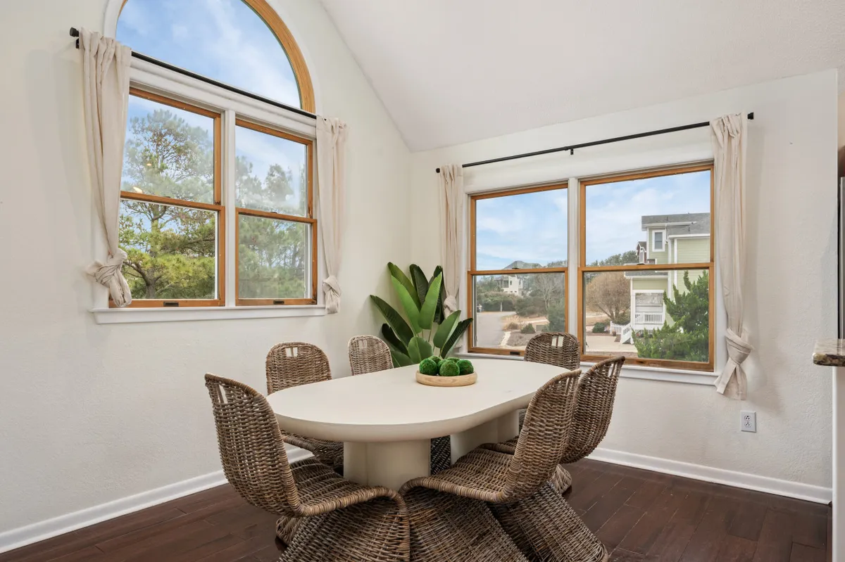 A warm and welcoming dining area features a round table surrounded by woven chairs, set against a backdrop of large windows and a leafy indoor plant. The space is designed for group meals, with ample seating and natural light fostering a convivial, celebratory atmosphere.