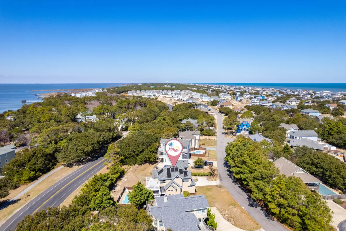 This image captures an expansive ocean view alongside a landmark building with a palm tree sign, a road leading to the beach, and part of the surrounding residential area. The scene conveys a sense of arrival and adventure, suggesting proximity to the beach, local shops, and the vibrant community.