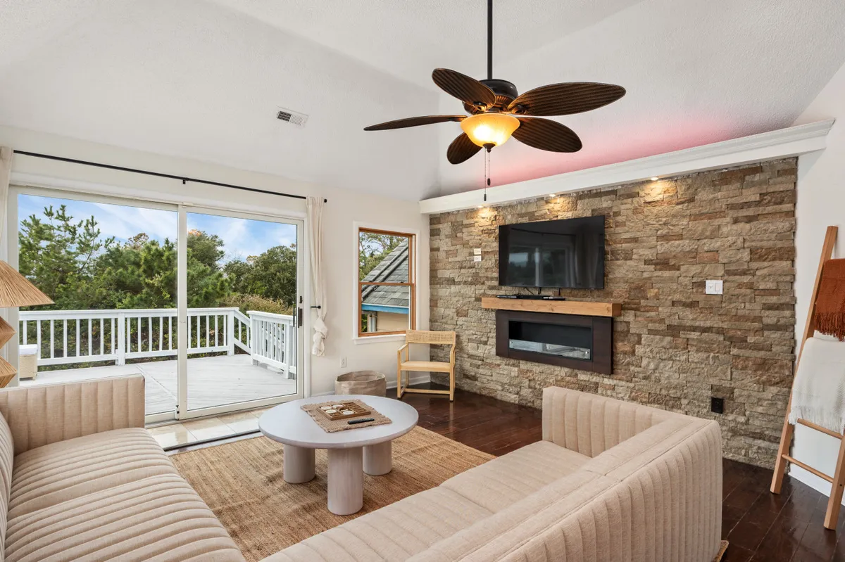 A welcoming living room with a plush sofa and coffee table facing a large TV mounted on a stone accent wall. Natural light pours in through a sliding glass door, which offers a glimpse of the deck outside, creating a bright yet cozy atmosphere.